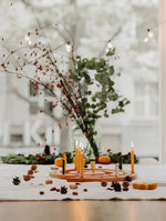 An autumnal tabletop arrangement with a Grimm's natural spiral celebration ring holding several lit yellow taper candles, surrounded by pinecones, acorns, small pumpkins, and a glass vase with greenery and red berries in front of a softly blurred window.