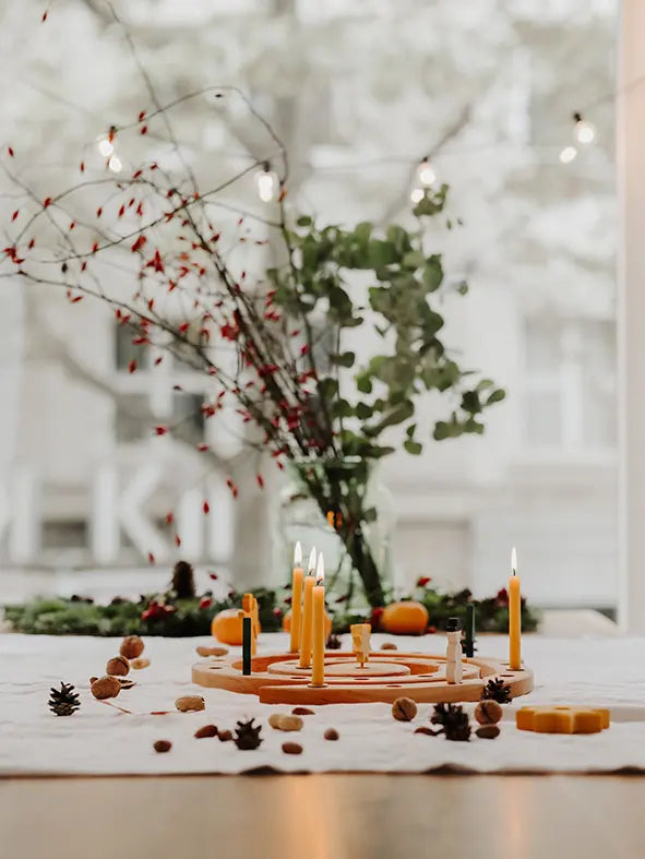An autumnal tabletop arrangement with a Grimm's natural spiral celebration ring holding several lit yellow taper candles, surrounded by pinecones, acorns, small pumpkins, and a glass vase with greenery and red berries in front of a softly blurred window.