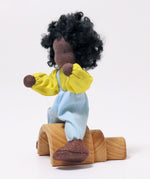 A side view of Grimm's Handmade Doll - Max sitting on a Grimm's small natural rainbow. The image is on a light grey background