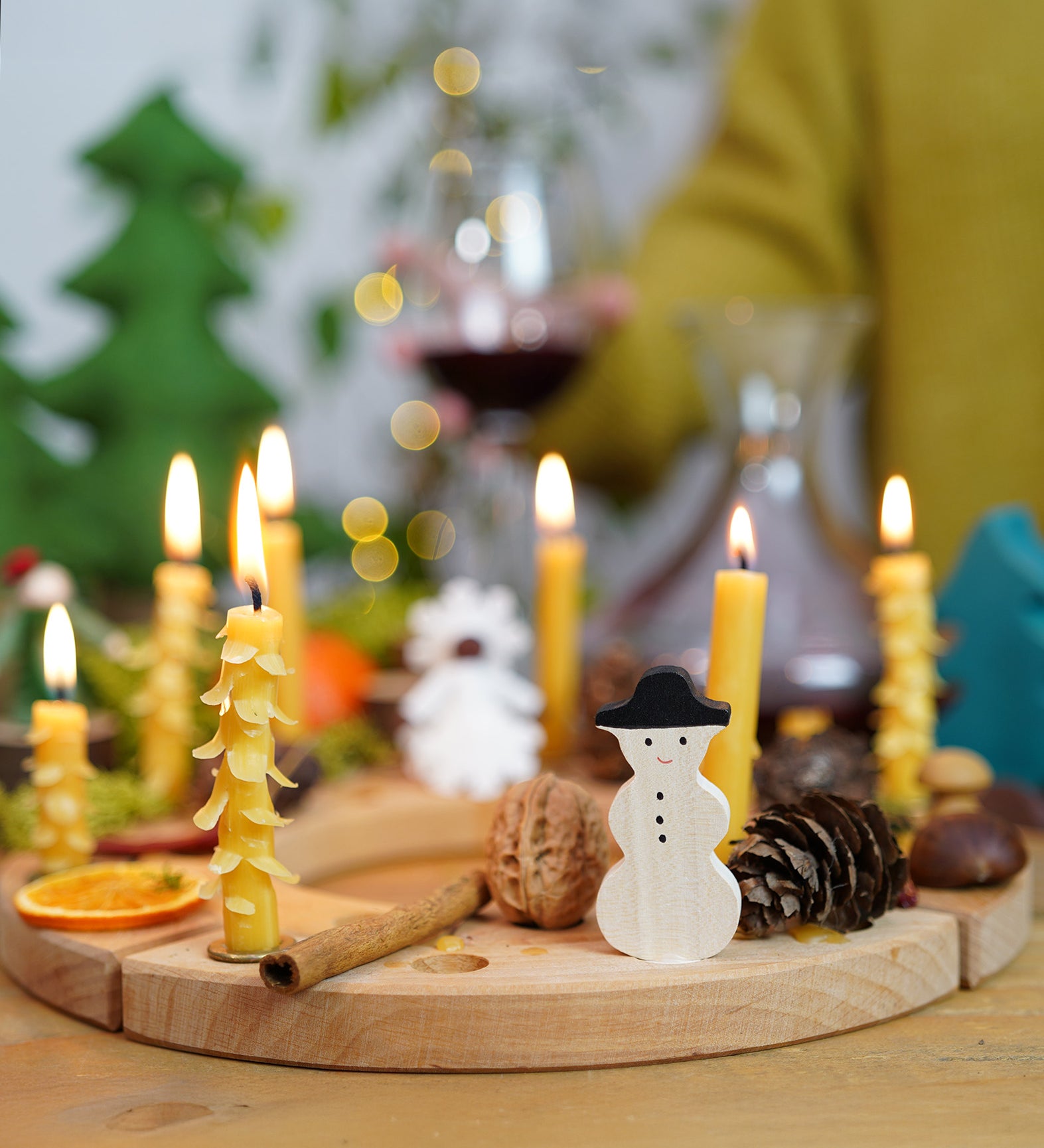 A Grimm's Wooden Celebration Ring, filled with Grimm's wooden Christmas decorative figures and candles in brass holders. There are also other decorative items such as a walnut, cinnamon stick, pine cone and conkers to create an uplifting winter display