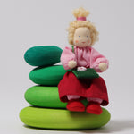The Grimm's Blonde Girl Doll, sitting down on the Grimm's Moss Pebbles Building Set, on a grey background