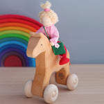 The Grimm's Blonde Girl Waldorf Doll is sat on a Grimm's Wooden Horse, with the Grimm's Rainbow 12 piece in the background