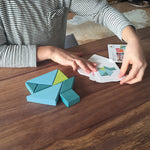 A child is playing with the Grimm's Tangram toy on a dark brown table, creating the puzzles using puzzle cards