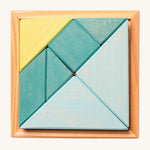 A wooden Tangram puzzle in shades of blue and green sits inside a small light wooden base on a cream background. There are 7 puzzle pieces in different shapes.