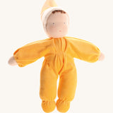 Grimm's Yellow Soft Doll is a hand-crafted Waldorf doll, wearing a yellow all-in-one baby grow with a matching night cap, with a smiling face and brown curls peeking out from under the yellow cap. The image is on a cream background