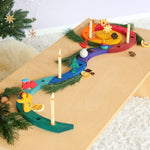 A colourful Grimm's wooden Advent spiral celebration ring with carved slots and small decorative pieces, shown on a light wood table with lit candles, evergreen branches, dried orange slices, and small holiday ornaments in a cozy indoor setting.