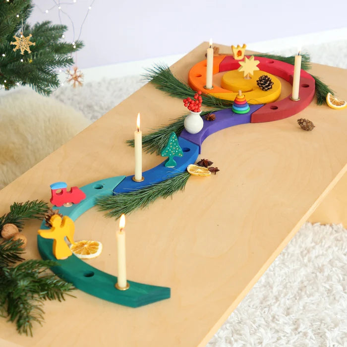 A colourful Grimm's wooden Advent spiral celebration ring with carved slots and small decorative pieces, shown on a light wood table with lit candles, evergreen branches, dried orange slices, and small holiday ornaments in a cozy indoor setting.