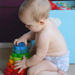 A child is sat down on the floor, infornt of a light blue toy box and is playing with the Grimm's Wooden Stacking Flower Toy. The child is holding the side of the flower and is attempting the take off the top flower block.