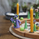 A beautiful Ambrosius doll sits on a Grimm's Wooden celebration ring, with yellow candles and green loose parts, to create a vibrant spring celebration display