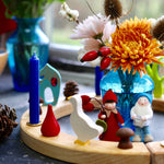 A Grimm's Wooden Celebration ring is used in a pretty winter display using Grimm's candles, Grimm's decorative figures and other loose parts. In the centre is a blue vase with flowers, and two Ambrosius dolls