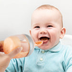 A baby smiling and eating from the Generation 3 Silicone Bottle Feeding Spoon Head, as their parent holds the feeding spoon to the babies mouth