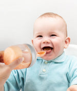 A baby smiling and eating from the Generation 3 Silicone Bottle Feeding Spoon Head, as their parent holds the feeding spoon to the babies mouth