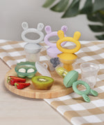A selection of Haakaa Freeze-N-Feed Mini Sets in different colours, set on a light wooden table with table cloth. Some of the feeders have food inside 
