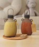 Two Haakaa Happii Bear Silicone Goody Pouches on a table with a label attached to the bear ear on the bottle. The left pouch is filled with yellow liquid, and the right is filled with orange/red liquid