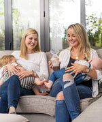Two people sat in a living room, breast feeding their children with one holding the Generation 3 160ml Breast Pump in peach