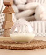 The Haakaa Ladybug Silicone Breast Milk Collector, filled with milk is stood on a light brown table and is stable due to the little silicone feet on the bottom of the milk collector
