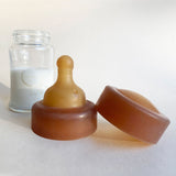 A Hevea Glass Baby Bottle with milk in the background with the natural rubber teat and cap in the foreground. 