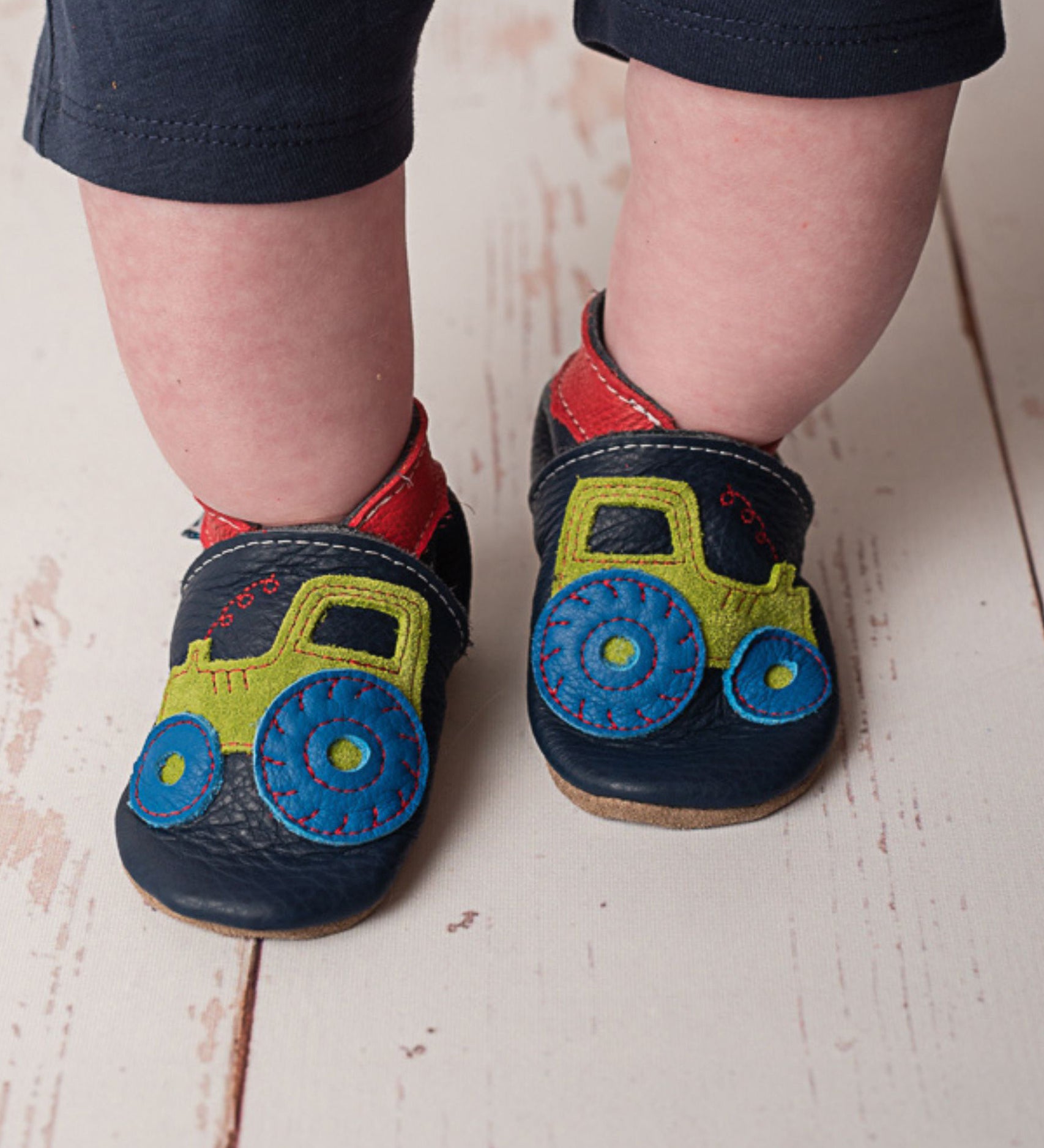A close up of a child wearing the Inch blue navy tractor baby shoe available at Babipur showing the red ankle cuff detail and the playful green suede tractor design