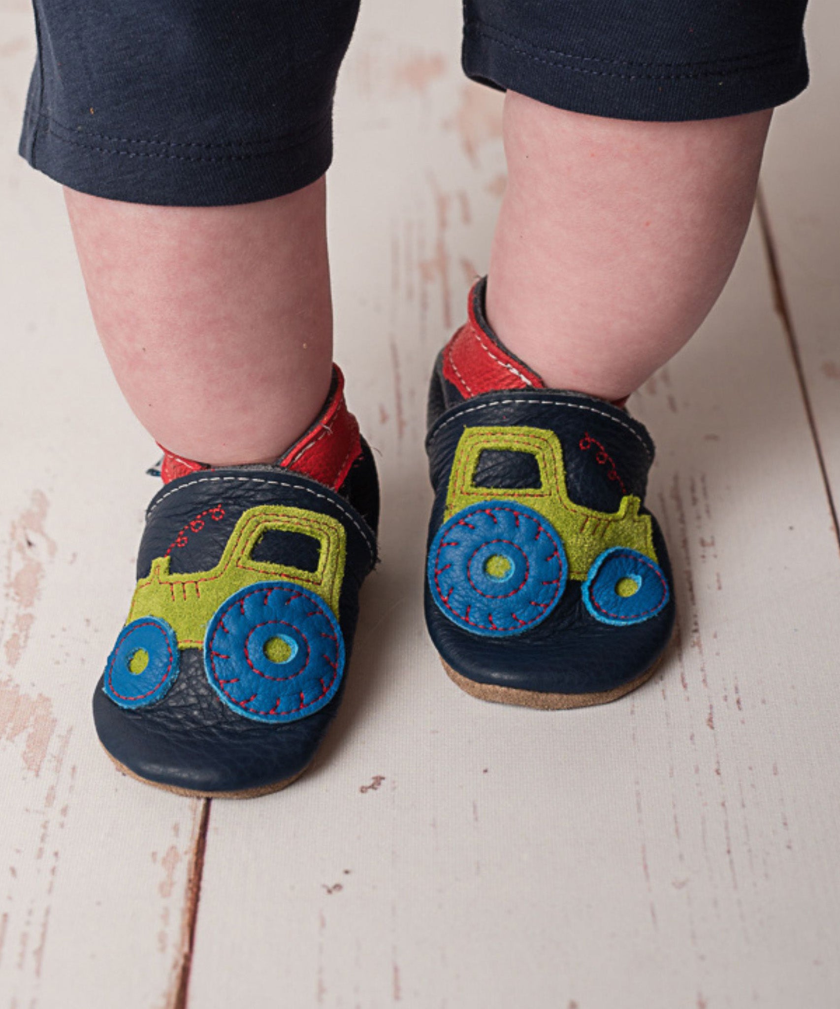 A close up of a child wearing the Inch blue navy tractor baby shoe available at Babipur showing the red ankle cuff detail and the playful green suede tractor design