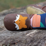 A close up of a child sitting on a wooden surface and wearing the Inch blue chocolate Mr Fox baby shoe available at Babipur showing the cute playful suede fox design