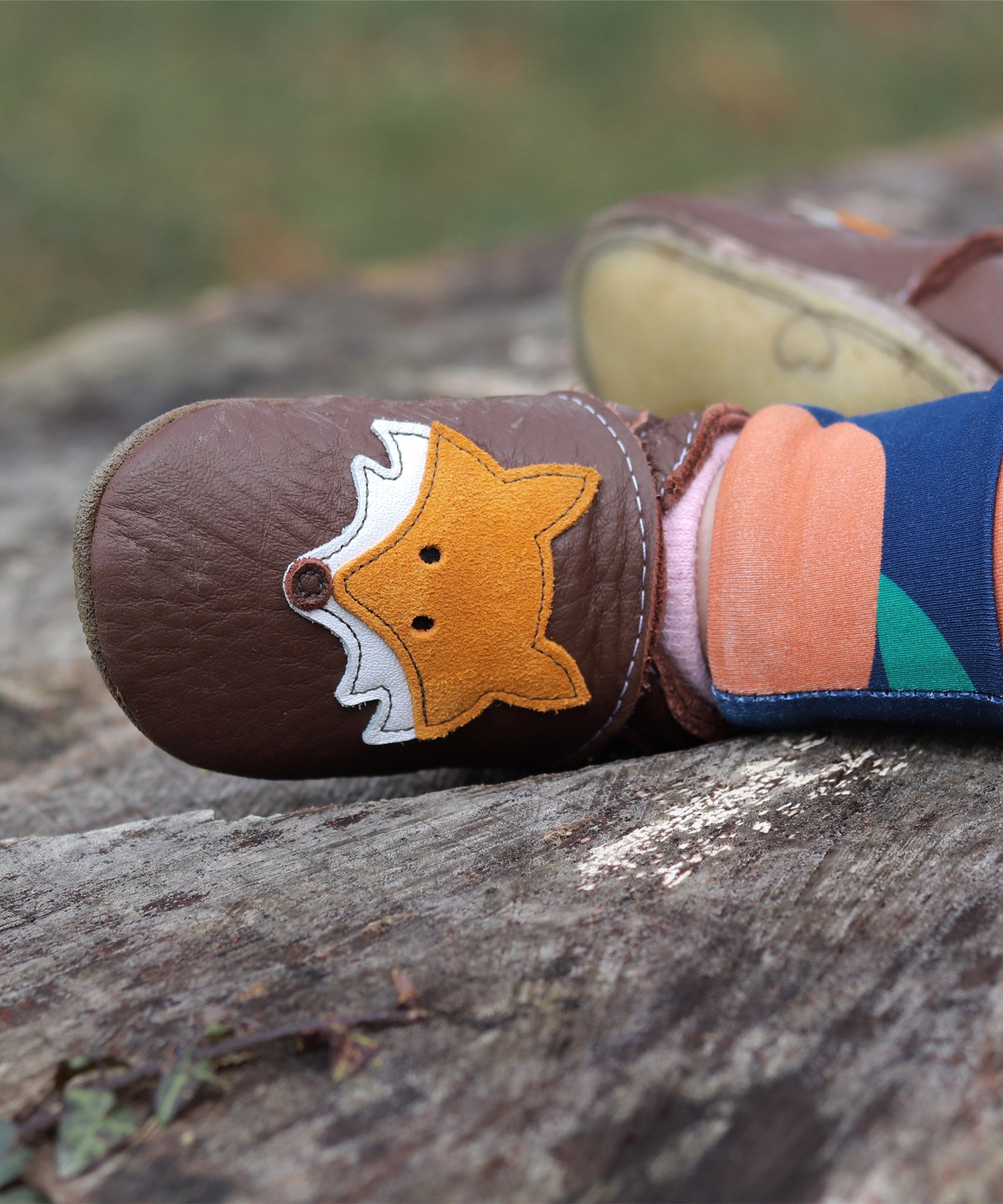 A close up of a child sitting on a wooden surface and wearing the Inch blue chocolate Mr Fox baby shoe available at Babipur showing the cute playful suede fox design