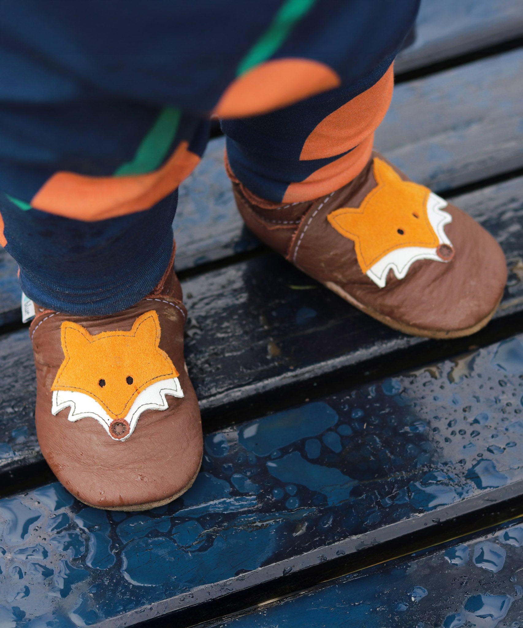 A close up of a baby standing wooden bench and wearing the Inch blue chocolate Mr Fox baby shoe showing the detail on the cute suede fox 