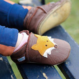A close up of a child sitting on a wooden bench and wearing the Inch blue chocolate Mr Fox baby shoe showing the stitching detail and a close up of the cute suede fox. 