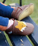 A close up of a child sitting on a wooden bench and wearing the Inch blue chocolate Mr Fox baby shoe showing the stitching detail and a close up of the cute suede fox. 