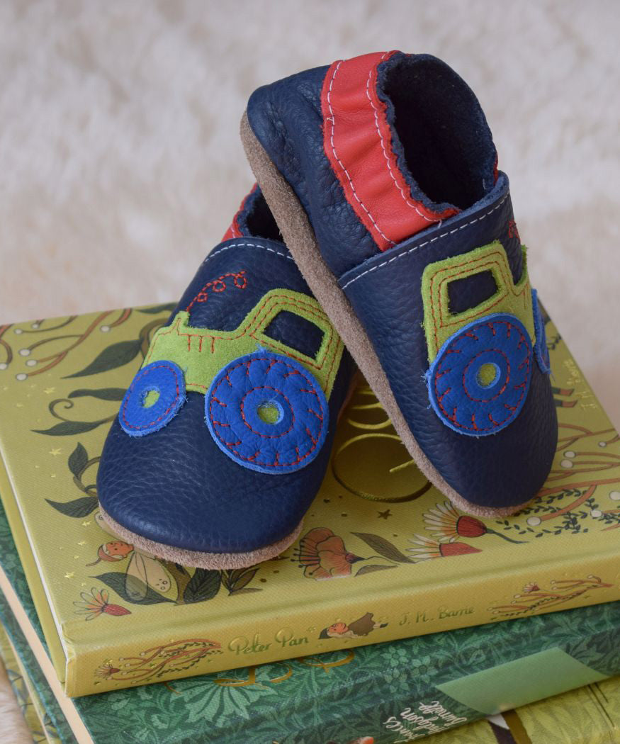 Inch Blue navy tractor baby shoes available at Babipur standing on top of children's story books. 