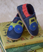 Inch Blue navy tractor baby shoes available at Babipur standing on top of children's story books. 