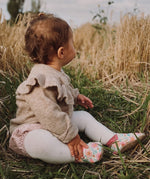 A baby sitting on grass wearing the Inch Blue wildroses shoes available at Babipur. 