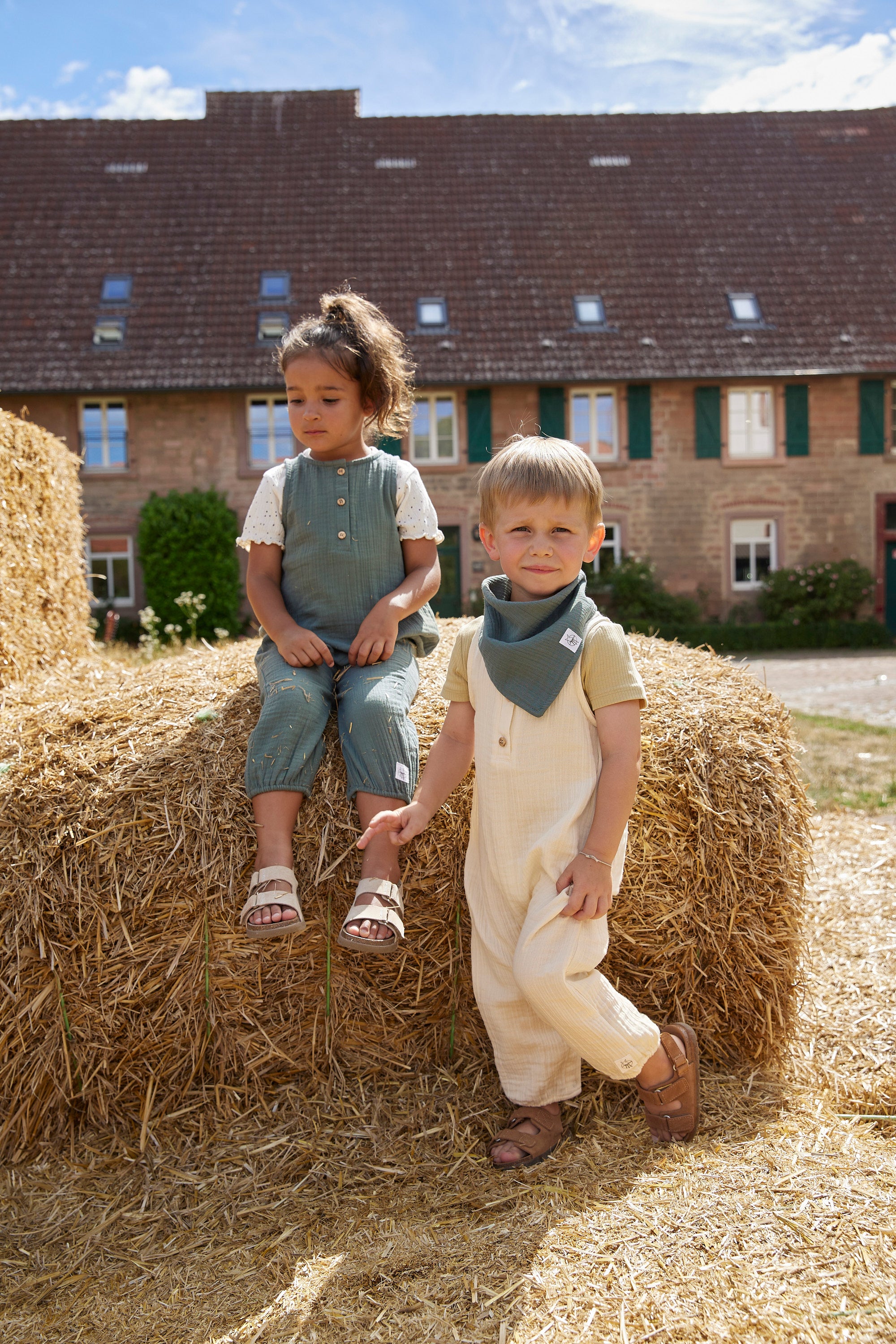 Two children sat on haybales, wearing Lassig organic cotton muslin jumpsuits. 