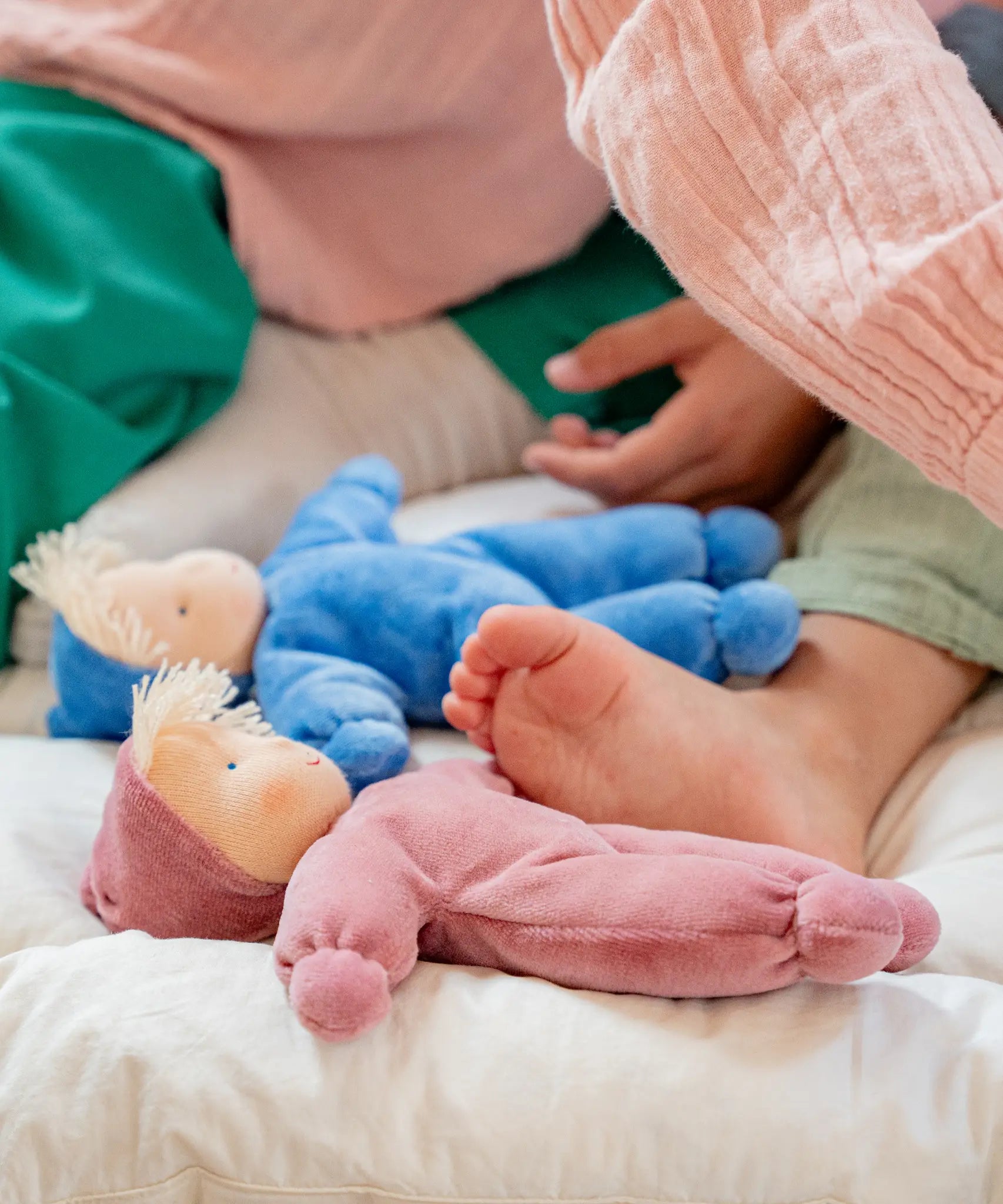 Children playing on a white blanket with a Kathe Kruse Lolo Rose waldorf doll lying flat next to a blue doll