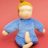 Kathe Kruse cloud nana waldorf doll showing a blue outfit and mustard yellow knitted hat sitting infront of a red background