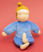 Kathe Kruse cloud nana waldorf doll showing a blue outfit and mustard yellow knitted hat sitting infront of a red background