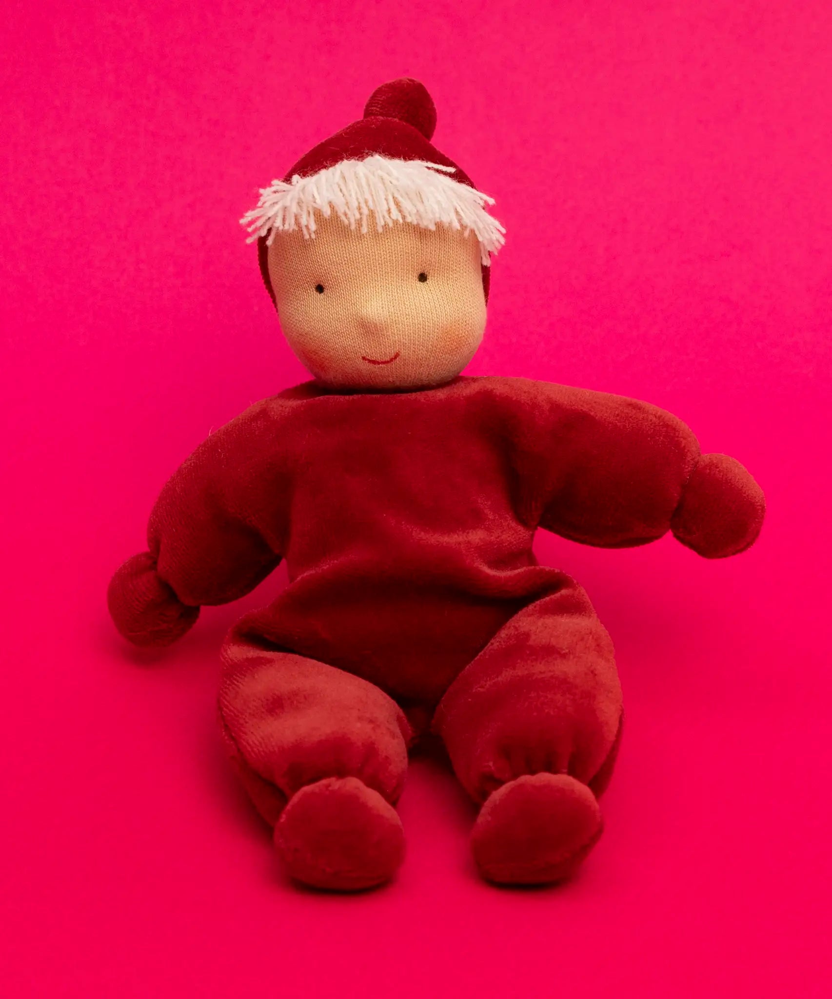 Kathe Kruse Lolo Ruby Waldorf doll showing a red towelling outfit sitting in front of a bright red background.