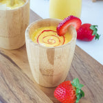 A yellow mango smoothie with red strawberry puree swirl in a bamboo baby cup, decorated with strawberry slices..