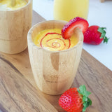 A yellow mango smoothie with red strawberry puree swirl in a bamboo baby cup, decorated with strawberry slices..