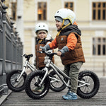 Two children happily stood with their Early Rider Big Foot Balance Bikes next to a railing, with a large light yellow building in the background