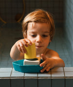 A child playing with the Oli & Carol Boat Bath Toy - Yellow, on the side of a blue tiled bath