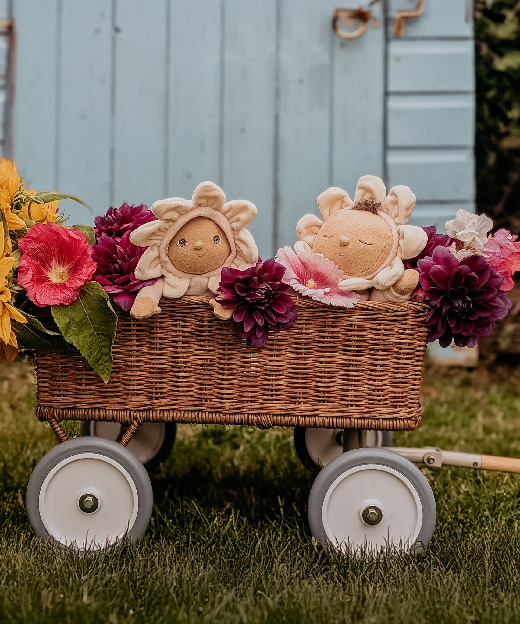 The Olli Ella Limited Edition Blossom Buds Gift Set - Gardenia, showing the Tootsie Gardenia & Dinky Dinkum Doll - Gracie Gardenia sat inside an Olli Ella cart with beautiful purple and pink flowers