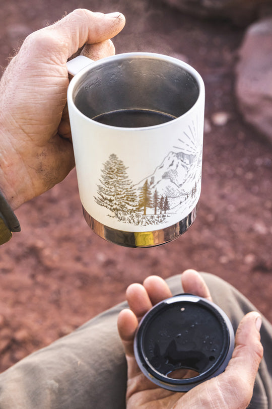 Hands holding a Klean Kanteen white insulated camp mug with a mountain design on it, filled with steaming hot coffee, and a lid in the other hand, to represent Klean Kanteen sale at Babipur, official UK stockist.