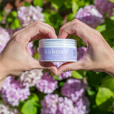 An adult hand holding the Kokoso 168g baby organic coconut oil tub in a shape of a heart with flowers in the background. Tub available at Babipur.
