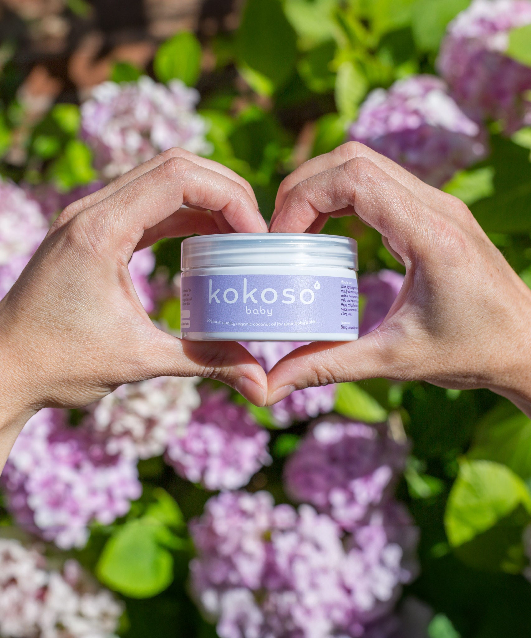 An adult hand holding the Kokoso 168g baby organic coconut oil tub in a shape of a heart with flowers in the background. Tub available at Babipur.