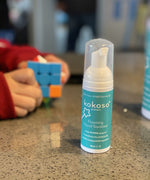 Kokoso protect foaming hand sanitiser original 50ml bottle available at Babipur on a stone surface with a child playing a puzzle game in the background.