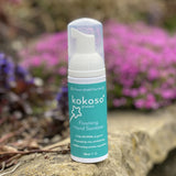 A close up of the Kokoso protect foaming hand sanitiser original 50ml bottle standing on a stone surface with flowers in the background. Sanitiser available at Babipur.