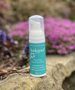 A close up of the Kokoso protect foaming hand sanitiser original 50ml bottle standing on a stone surface with flowers in the background. Sanitiser available at Babipur.