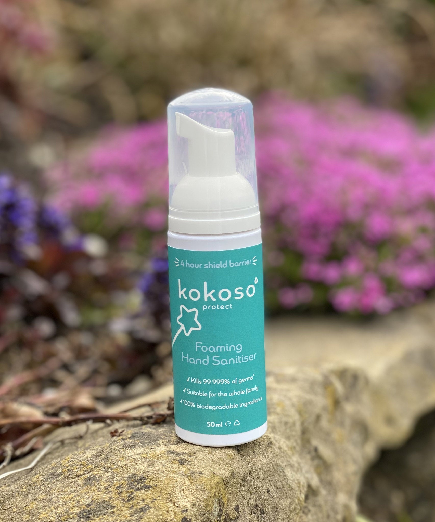 A close up of the Kokoso protect foaming hand sanitiser original 50ml bottle standing on a stone surface with flowers in the background. Sanitiser available at Babipur.