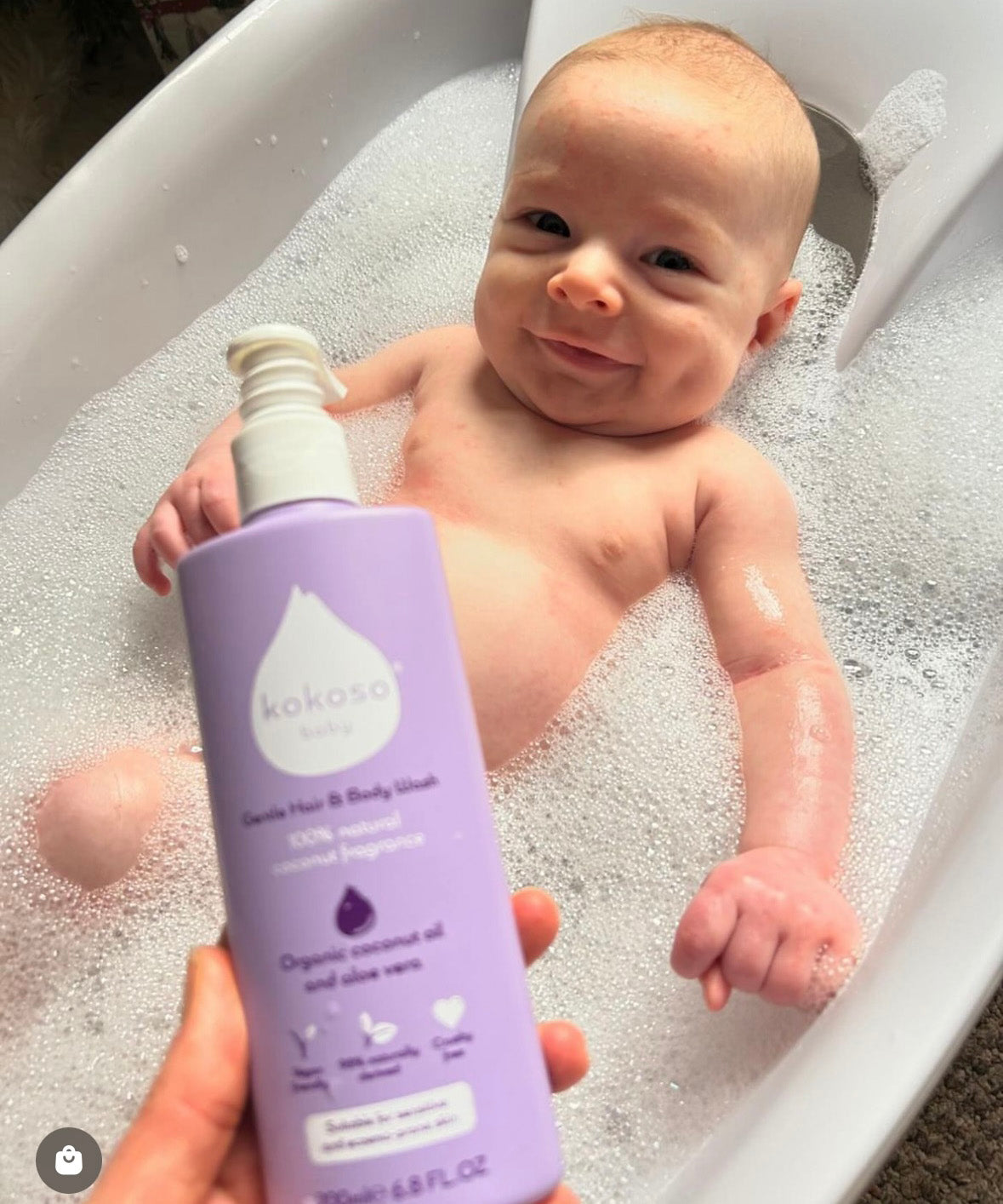A adult hand holding the Kokoso softly coconut scented organic baby body and hair wash 200ml bottle above a baby in a bath. Bottle available at Babipur.