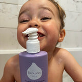 A close up of a child in a bath and holding the Kokoso softly scented organic baby hair body & wash bottle. Bottle available at Babipur.